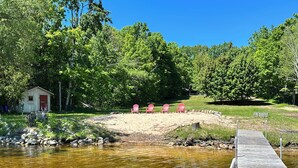 A wooden dock extending into a calm body of water, surrounded by lush greenery and a small shed with a red door. - Private beachfront cottage,  WiFi and AC on Lake Paradise, minutes from Mackinaw (Carp Lake)