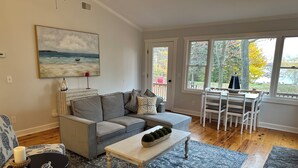 A living room with a gray sofa, a patterned armchair, a coffee table, and a painting of a beach scene. - Private beachfront cottage,  WiFi and AC on Lake Paradise, minutes from Mackinaw (Carp Lake)