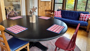 A wooden interior with a round table, chairs, a blue sofa, and a view of trees through large windows. - Private beachfront cottage,  WiFi and AC on Lake Paradise, minutes from Mackinaw (Carp Lake)
