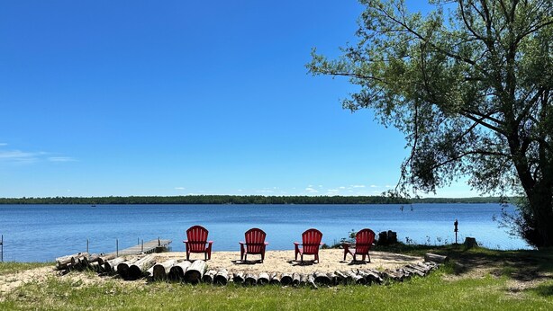 A lakeside view with four red Adirondack chairs arranged on a sandy area, a dock with a boat, and a tree to the right. - Private beachfront cottage,  WiFi and AC on Lake Paradise, minutes from Mackinaw (Carp Lake)