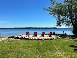 Blick auf einen See mit vier roten Adirondack-Stühlen, die auf einer sandigen Fläche angeordnet sind, einem Steg mit einem Boot und einem Baum auf der rechten Seite.