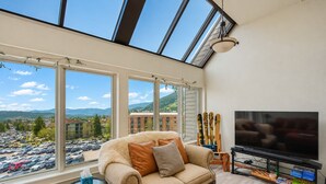 A living room with a sofa, TV, and ski equipment, large windows with a view of a parking lot and buildings. - Unique Park City Ski Resort Condos (Park City)