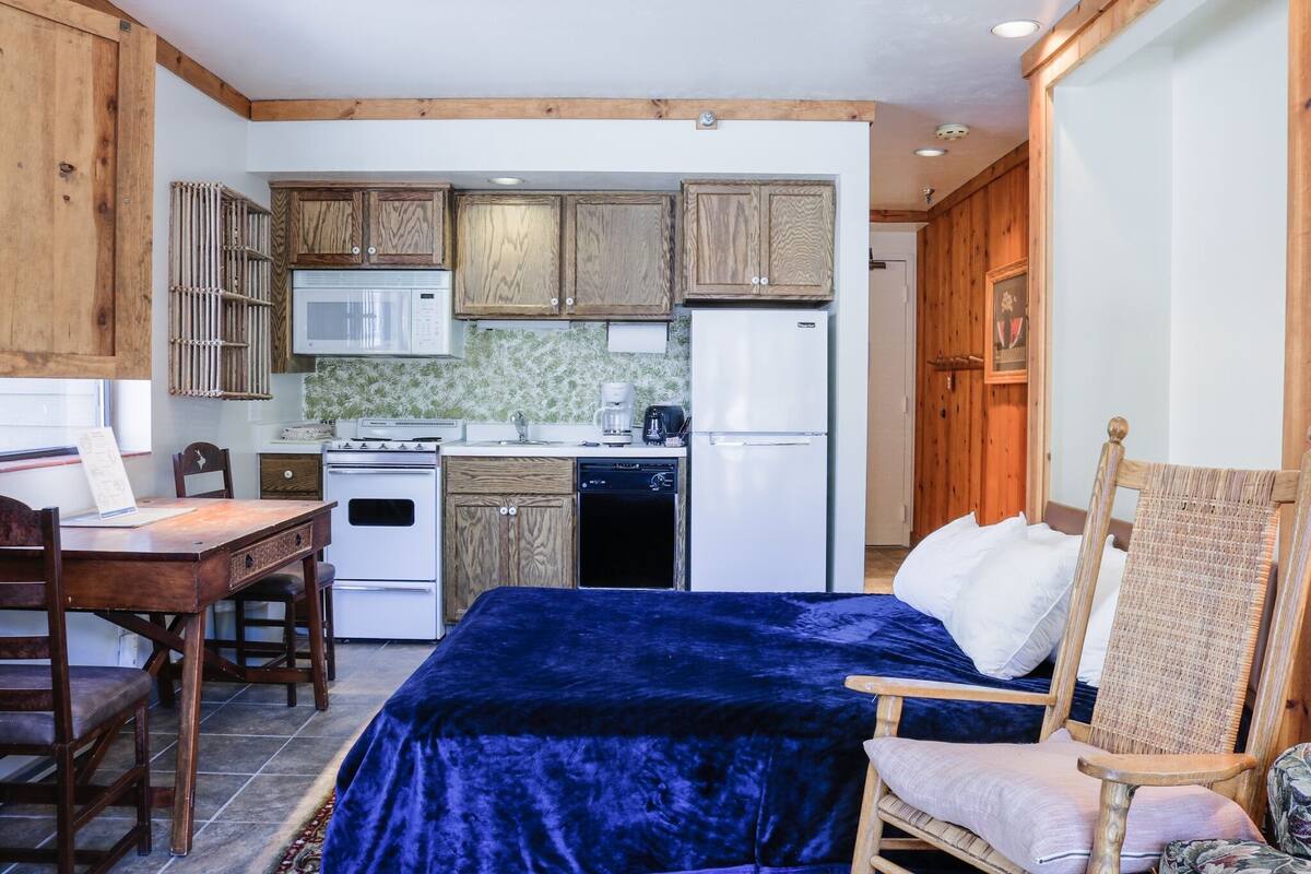 Ein kleines Zimmer mit Bett, Esstisch, Küchenzeile, Holzschränken, Mikrowelle, Kühlschrank und Kaffeemaschine.