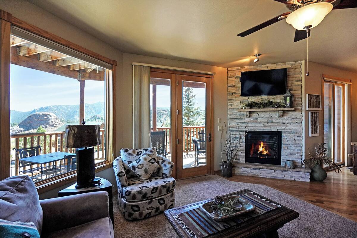 Un salon chaleureux avec une cheminée, un téléviseur fixé au-dessus, et un coin salon confortable offrant une vue sur les montagnes grâce à de grandes fenêtres.