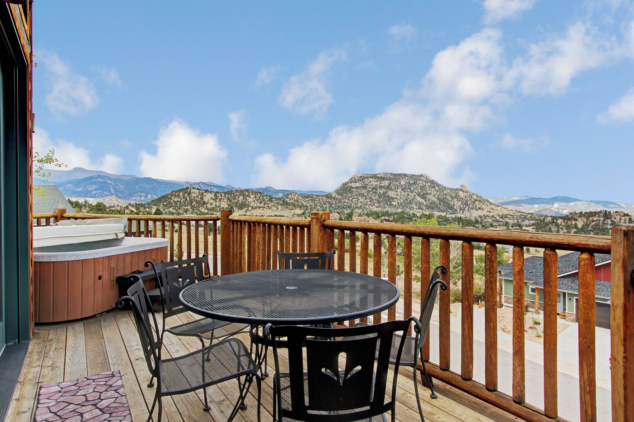 A wooden deck with a hot tub, round table, and chairs, overlooking a mountainous landscape.