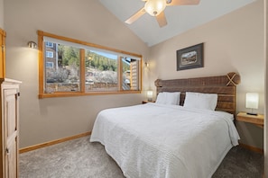 A bedroom with a large bed, a ceiling fan, a window with a view of trees and buildings, and a nightstand with a lamp.