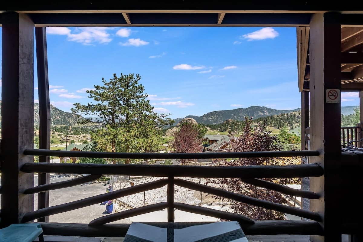 Une vue depuis une fenêtre ornée d’une rambarde en métal, donnant sur un paysage montagneux avec des arbres et un ciel dégagé.