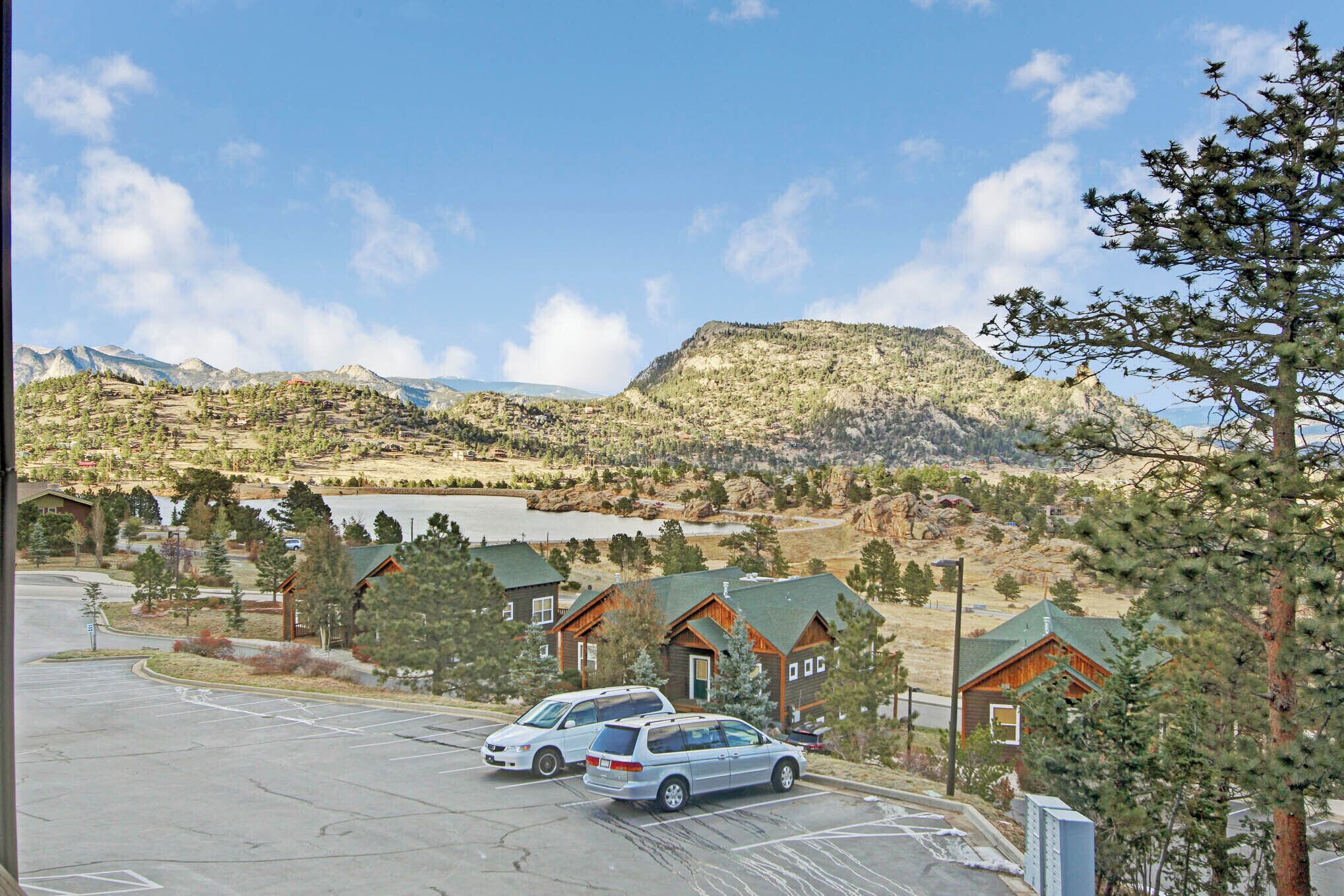 On peut voir un parking avec plusieurs véhicules, entouré de bâtiments aux façades en bois et aux toits végétalisés, le tout se détachant sur un paysage de montagnes et d’un lac.
