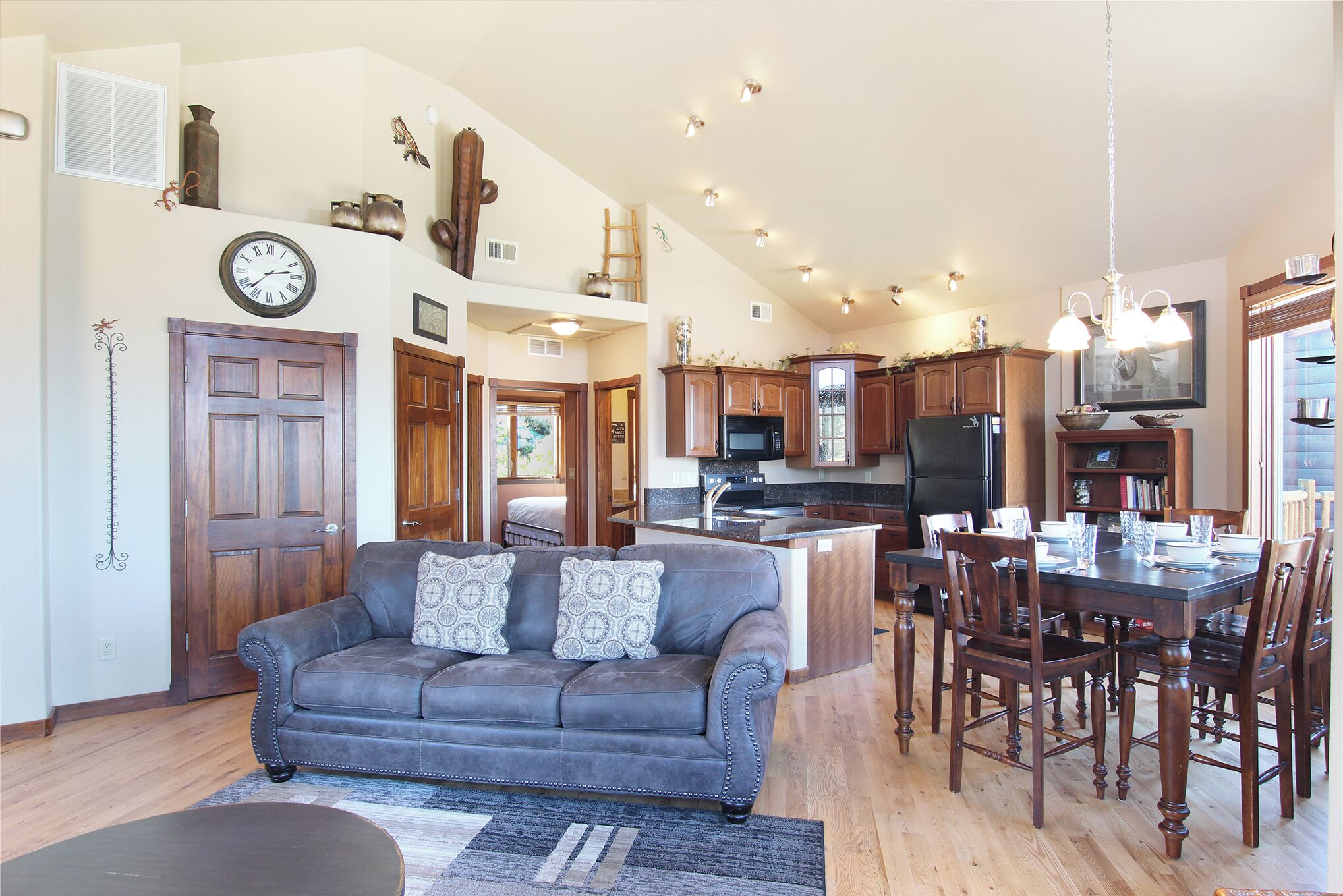 A spacious living area with a dark leather sofa, wooden dining table, and chairs, a kitchen with wooden cabinets, and a large clock on the wall.