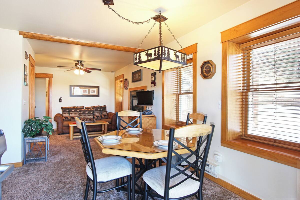 Un coin repas avec une table et des chaises en bois, un ventilateur de plafond et une grande fenêtre équipée de stores.