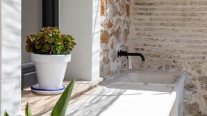 Ein weißes Waschbecken mit einem schwarzen Wasserhahn, ein Blumentopf mit grünem und rotem Blattwerk und eine Steinwand mit rauer Oberfläche.
