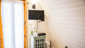 Une salle de bain avec une porte en bois, un rideau blanc, un petit meuble contenant des articles de toilette, des toilettes et un téléviseur fixé au mur.