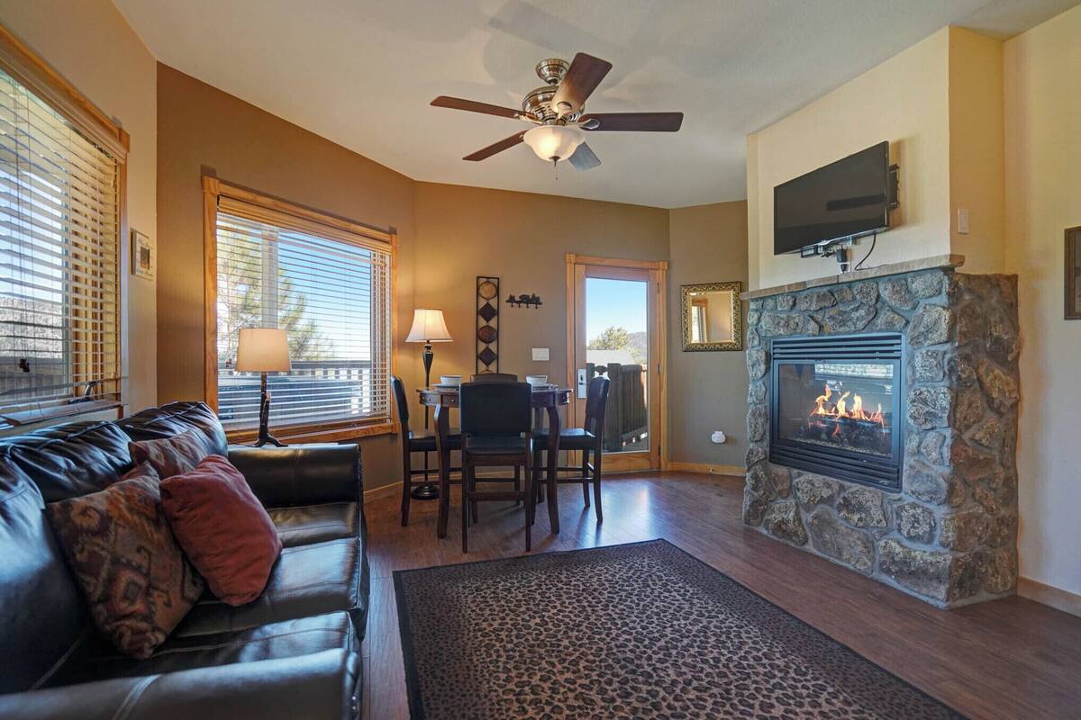 A living room with a fireplace, a ceiling fan, a television mounted above it, and a dining area with a table and chairs.
