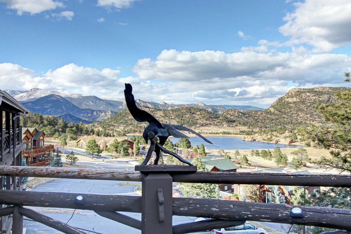 A bird sculpture on a railing overlooking a mountainous landscape with buildings and a lake.