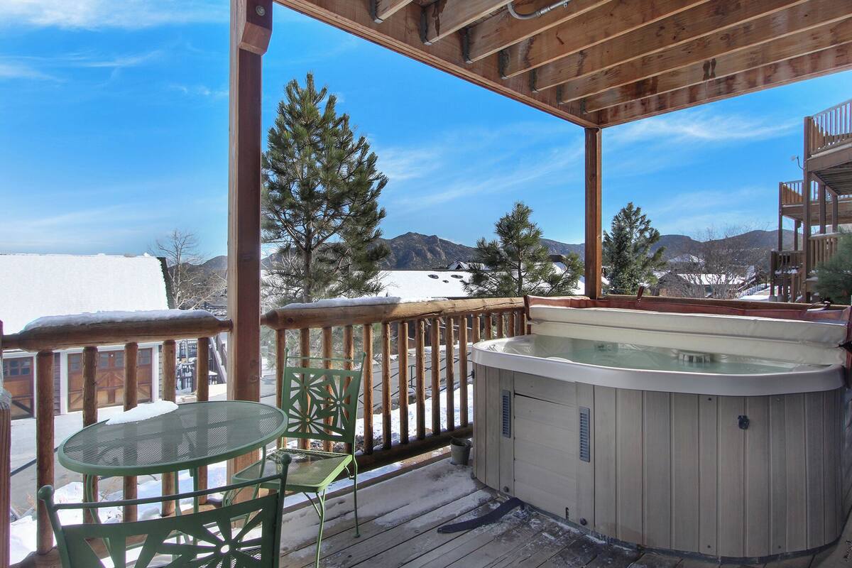 A wooden deck with a hot tub, a glass table, and a chair, overlooking a snowy landscape with mountains.