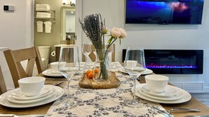 Een eetgedeelte met een tafel voor vier personen, een vaas met bloemen en een flatscreen televisie waarop een berglandschap te zien is.