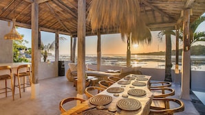 Un restaurante frente al mar con una larga mesa de madera dispuesta para comer, rodeado de sillas de mimbre, y vistas al océano.