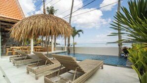 Zona de piscina con sillones de descanso, una sombrilla de paja y vista a la playa.