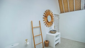 Un baño con inodoro blanco, escalera de madera, espejo redondo, lavamanos blanco y estante pequeño.