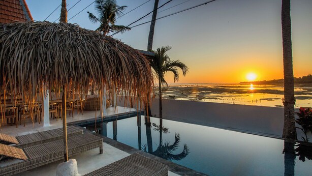 Una piscina infinita con sillones de descanso y una estructura de techo de paja, con vistas a una playa al atardecer.