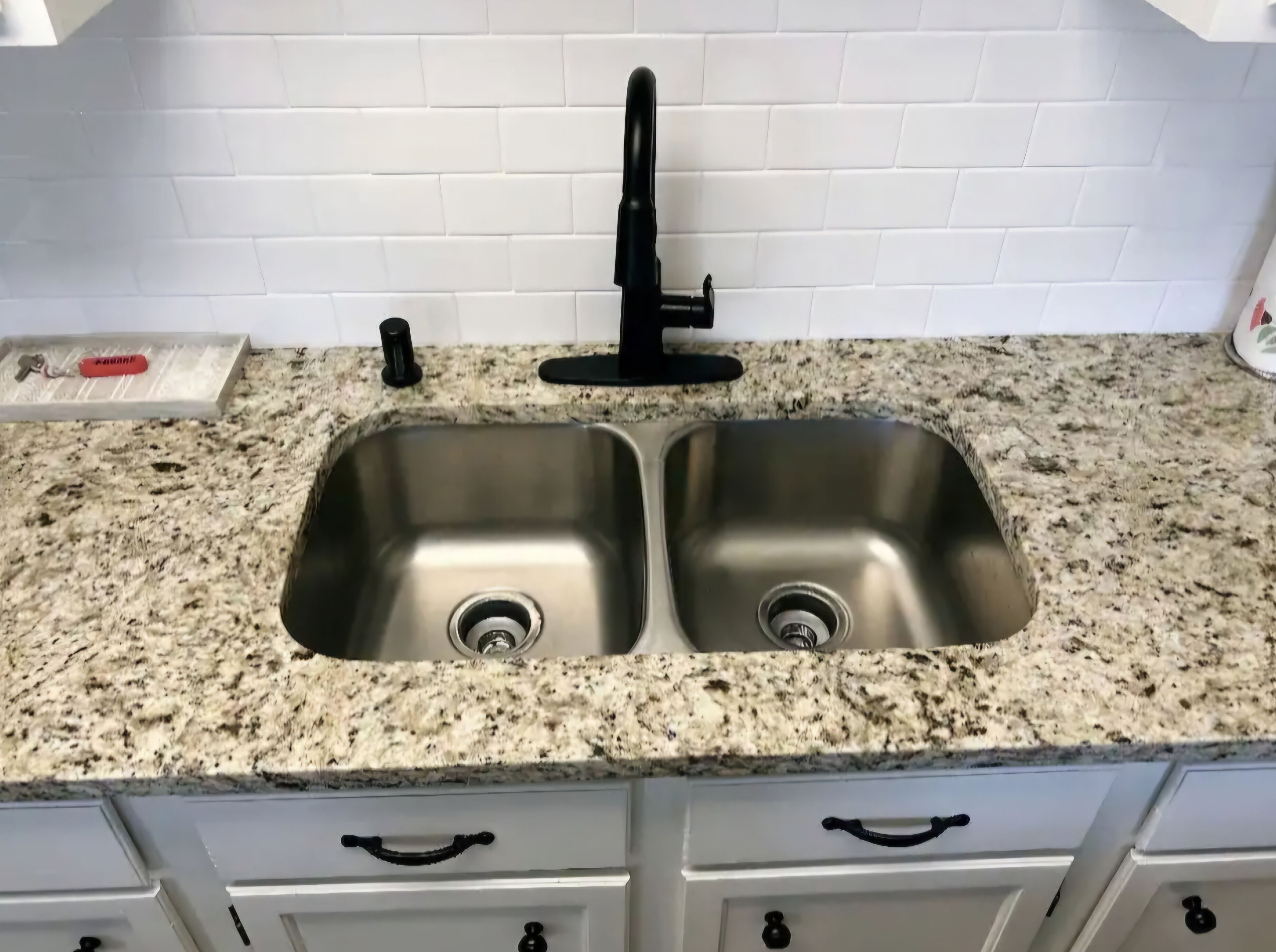 A kitchen with a double stainless steel sink, granite countertop, and white cabinetry.
