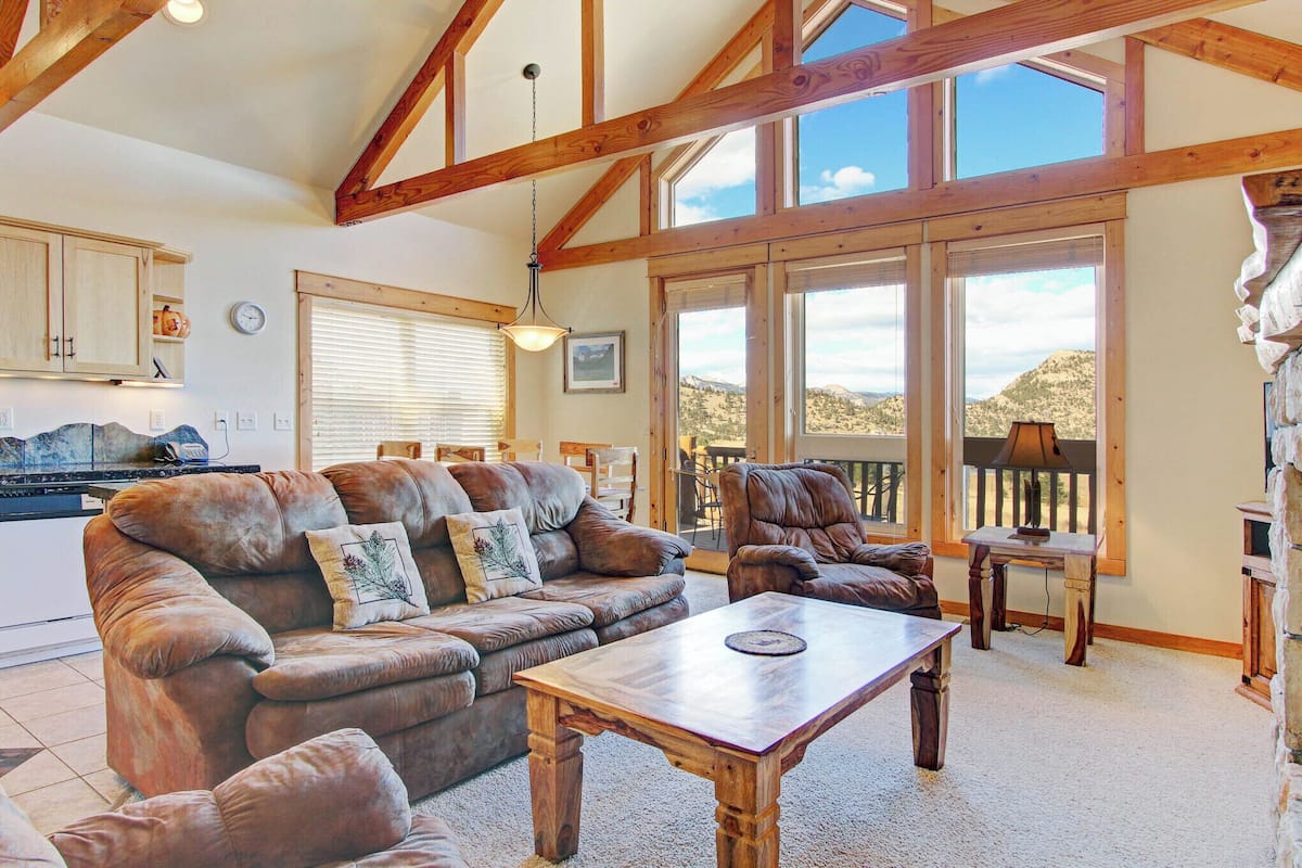 A spacious living room with a large wooden table, leather sofas, and a view of the mountains through large windows.