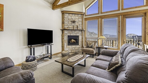 A living room with a stone fireplace, a flat-screen TV, and a large window with blinds. - Estes Park Vacation Lodging (Estes Park)