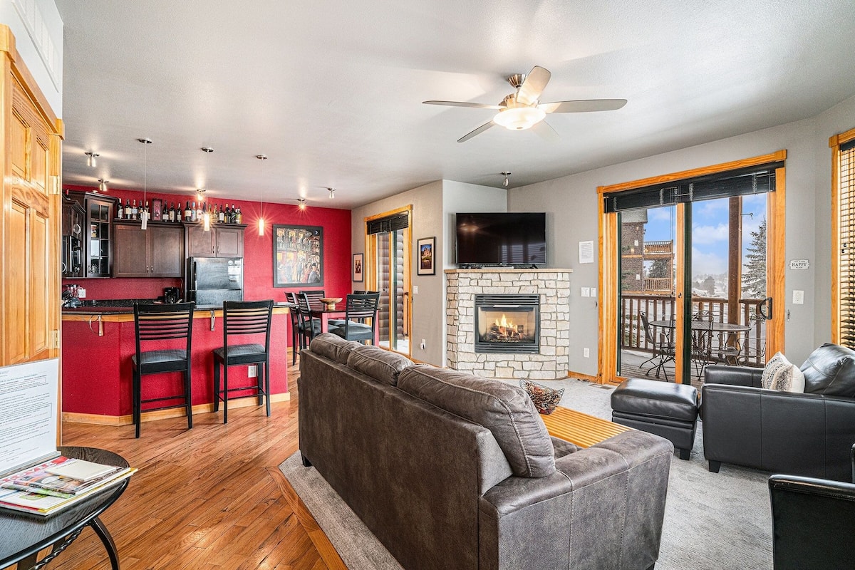 A spacious living area with a fireplace, a kitchen with red accents, and a balcony with a view.