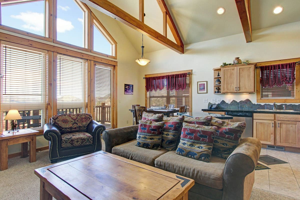 A cozy living room with a sofa, a wooden coffee table, and a dining area with a table and chairs. There are large windows with blinds, a kitchen area with cabinets, and a ceiling with exposed beams.