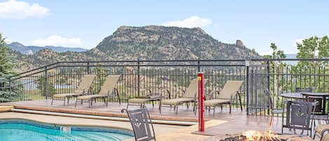 A pool area with lounge chairs and a fire pit, surrounded by a deck and a mountain view.