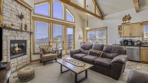 A spacious living room with a stone fireplace, large windows, a cozy sofa, and a kitchen area. - Estes Park Vacation Lodging (Estes Park)
