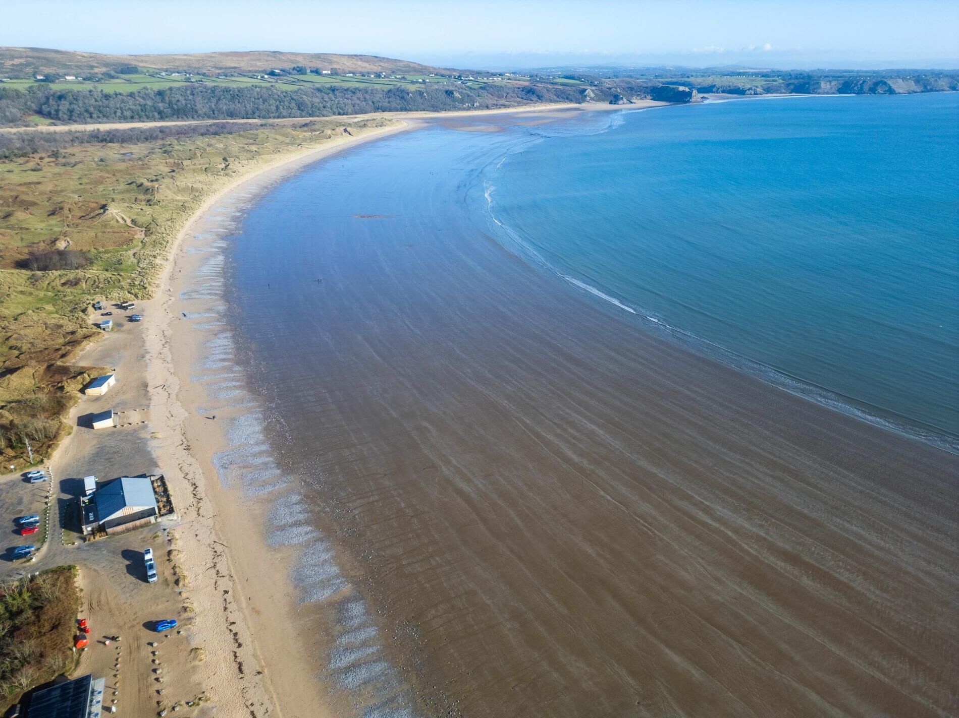 An aerial view of a coastal area with a sandy beach, a few buildings, and a clear view of the ocean.