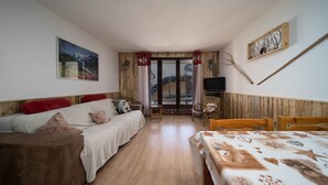 A cozy room with a wooden floor, a bed with a patterned duvet, a sofa with red cushions, a television on a wooden shelf, and a painting on the wall.