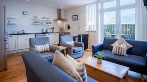 A modern living room with a kitchen, featuring a sofa set, a dining table, and a view of the outdoors.