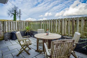 Eine hölzerne Terrasse im Freien mit einem runden Tisch, vier Stühlen, einer Weinflasche und einem Glas.