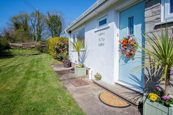 Een wit gebouw met een blauwe deur, een welkomstbord en een kleine tuin met bloempotten en bloemen.