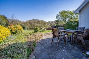 Een terras buiten met een houten tafel en stoelen, omringd door een tuin met verschillende planten en een tuinhuisje op de achtergrond.