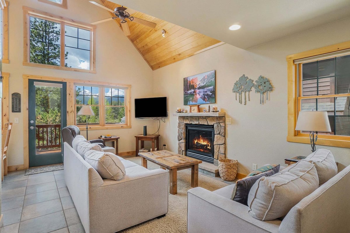 A cozy living room with a stone fireplace, comfortable sofas, a flat-screen TV, and large windows offering a view of the outdoors.