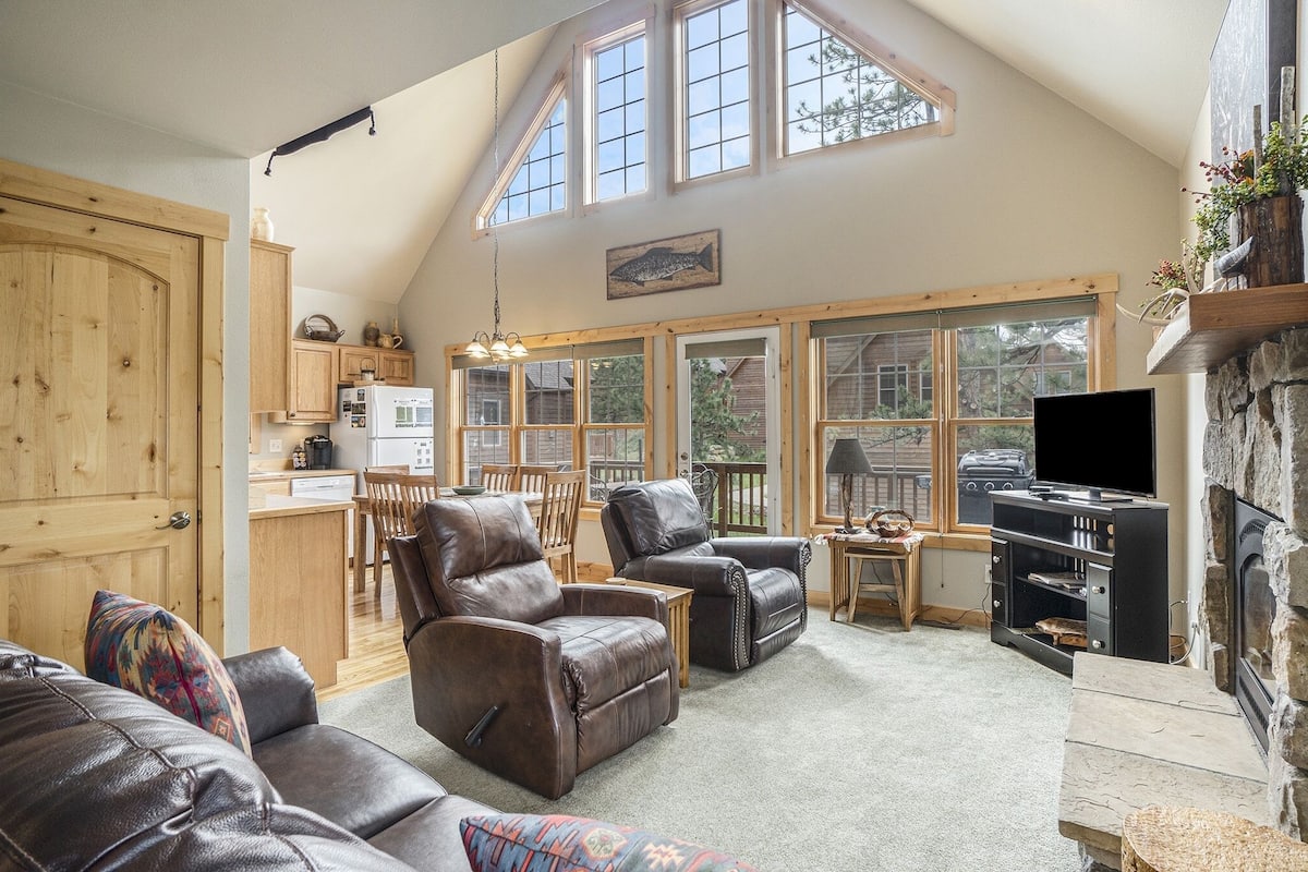 A spacious living room with leather sofas, a stone fireplace, and large windows offering views of the outdoors.