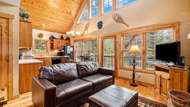 A cozy cabin interior with a wooden ceiling, a leather sofa, a dining area, and large windows offering a view of the forest.