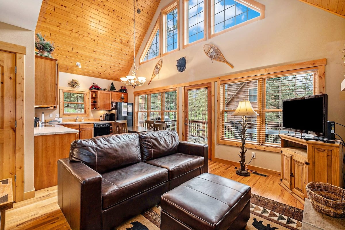 A cozy cabin interior with a wooden ceiling, a leather sofa, a dining area, and large windows offering a view of the forest.