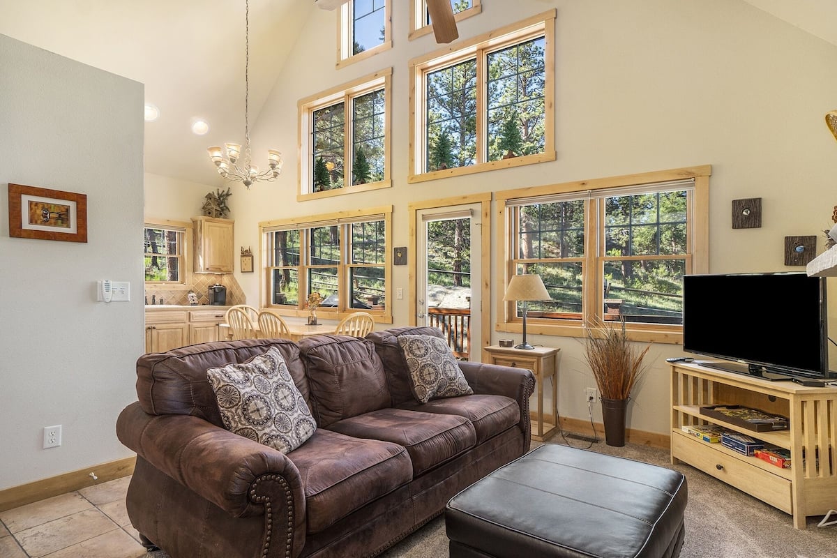 A spacious living room with a brown leather sofa, a black ottoman, and a wooden entertainment center with a flat-screen TV. Large windows provide views of the surrounding trees.