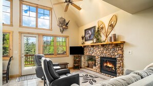 A living room with a stone fireplace, a ceiling fan, and a large window overlooking a deck.