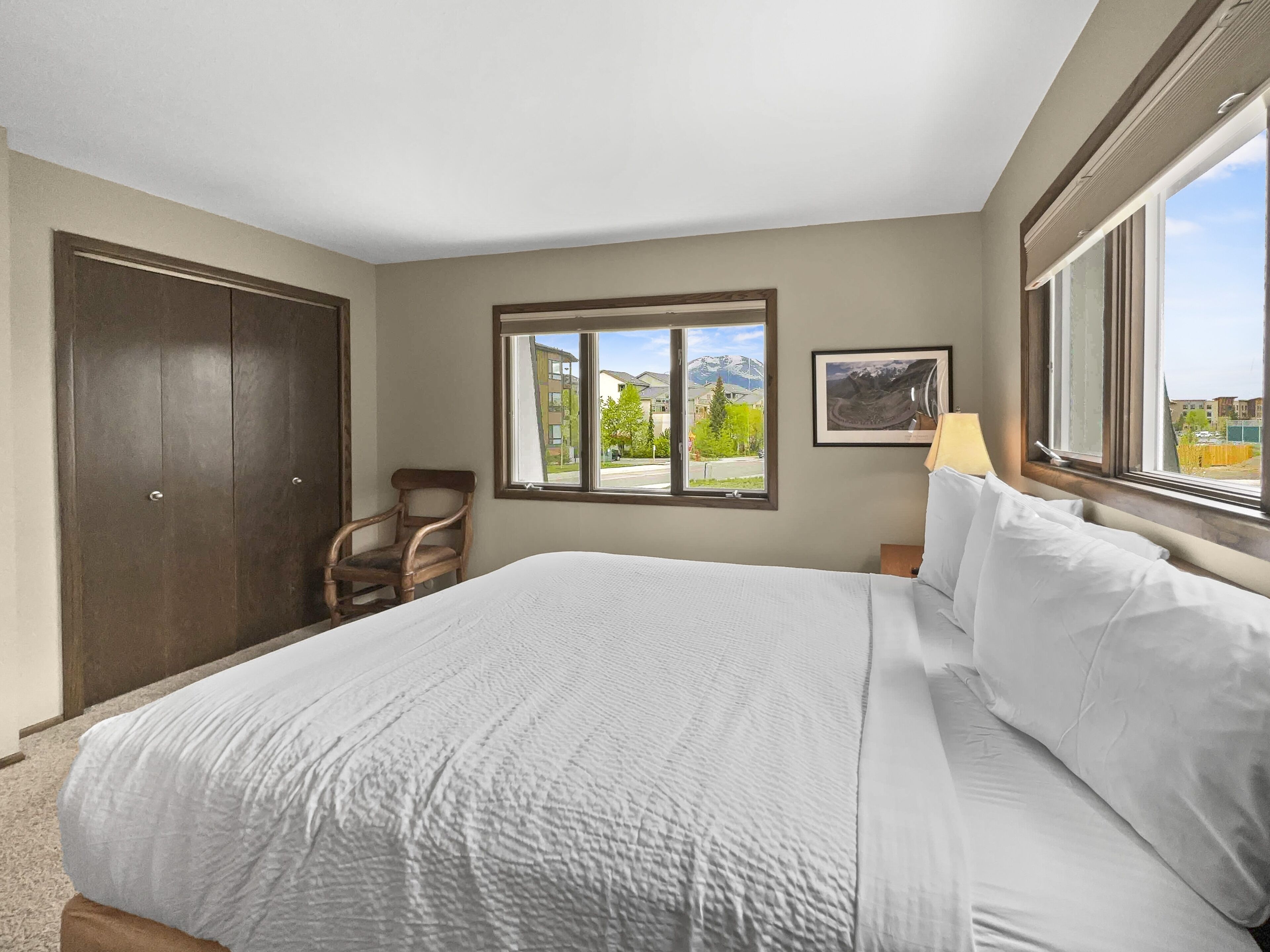 A hotel room with a large bed, a wooden chair, a window with a mountain view, and a framed picture on the wall.