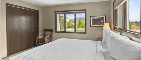 A hotel room with a large bed, a wooden chair, a window with a mountain view, and a framed picture on the wall.