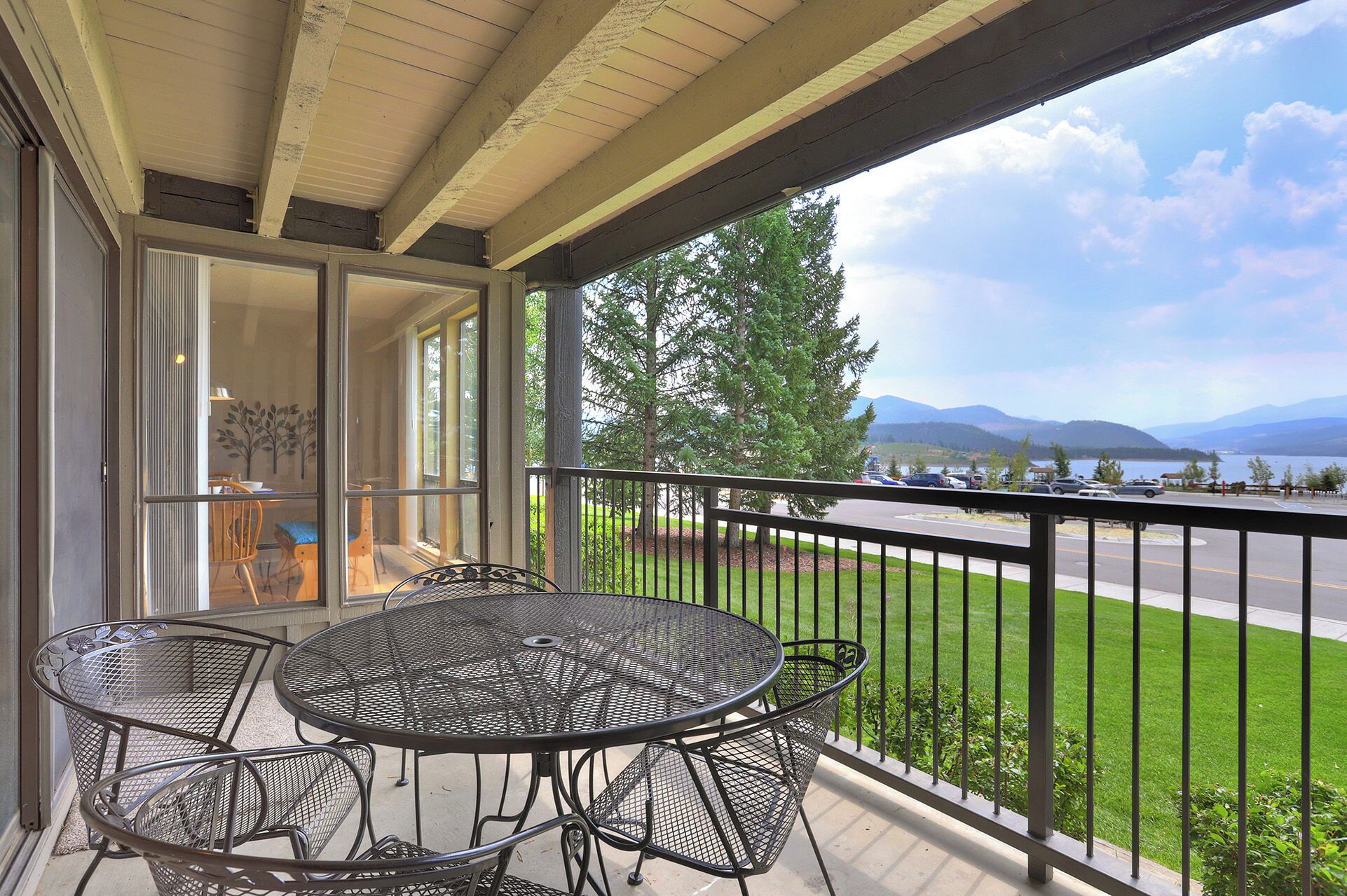 A balcony with a round metal table and four chairs, overlooking a parking area and a lake with mountains in the distance.
