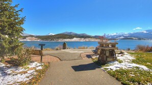 Ein gepflasterter Weg führt zu einer Bank und einer Steinstruktur am See, mit Bergen im Hintergrund.