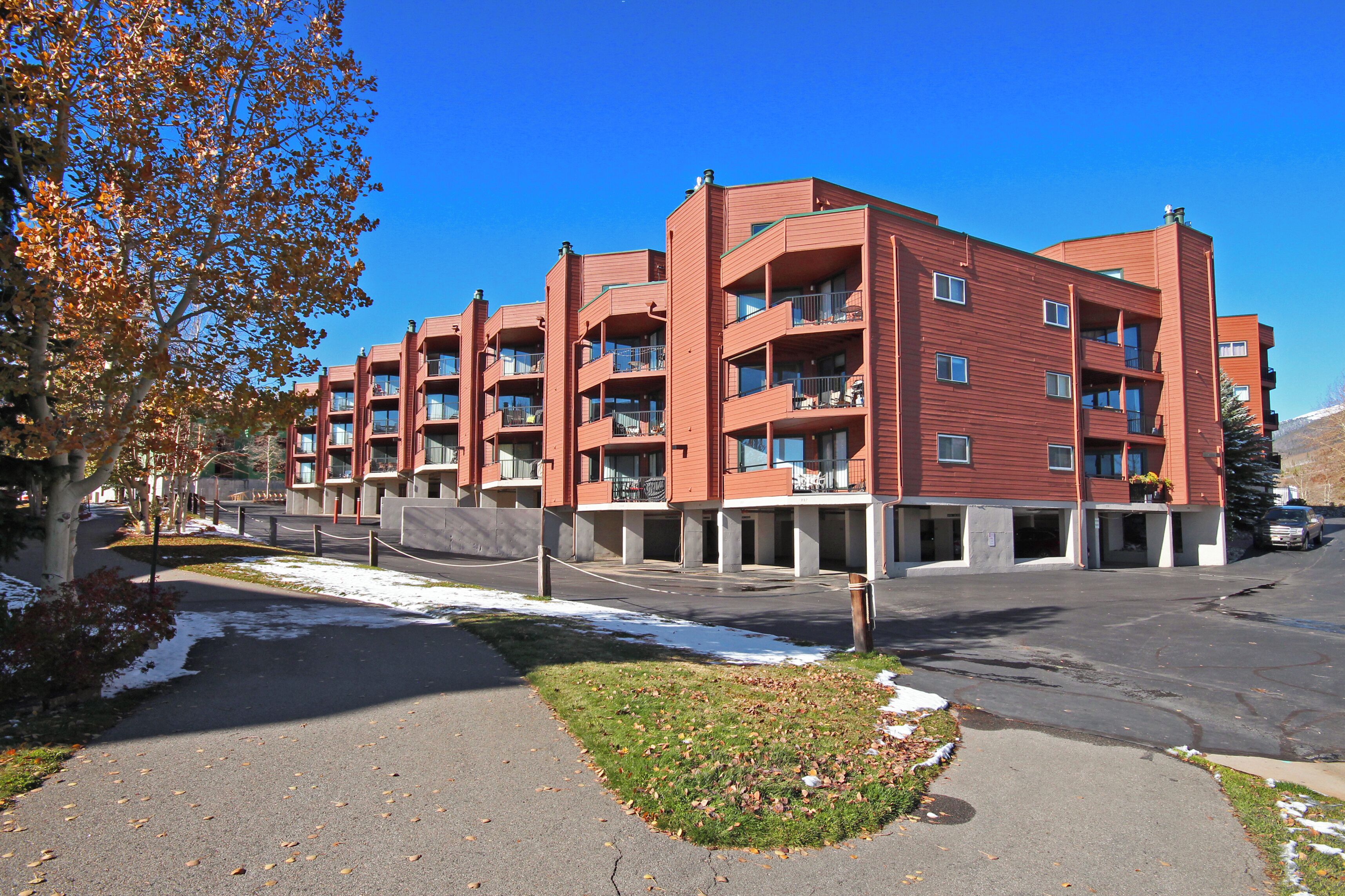 A multi-story red-brick building with balconies, a parking area, and a clear blue sky.