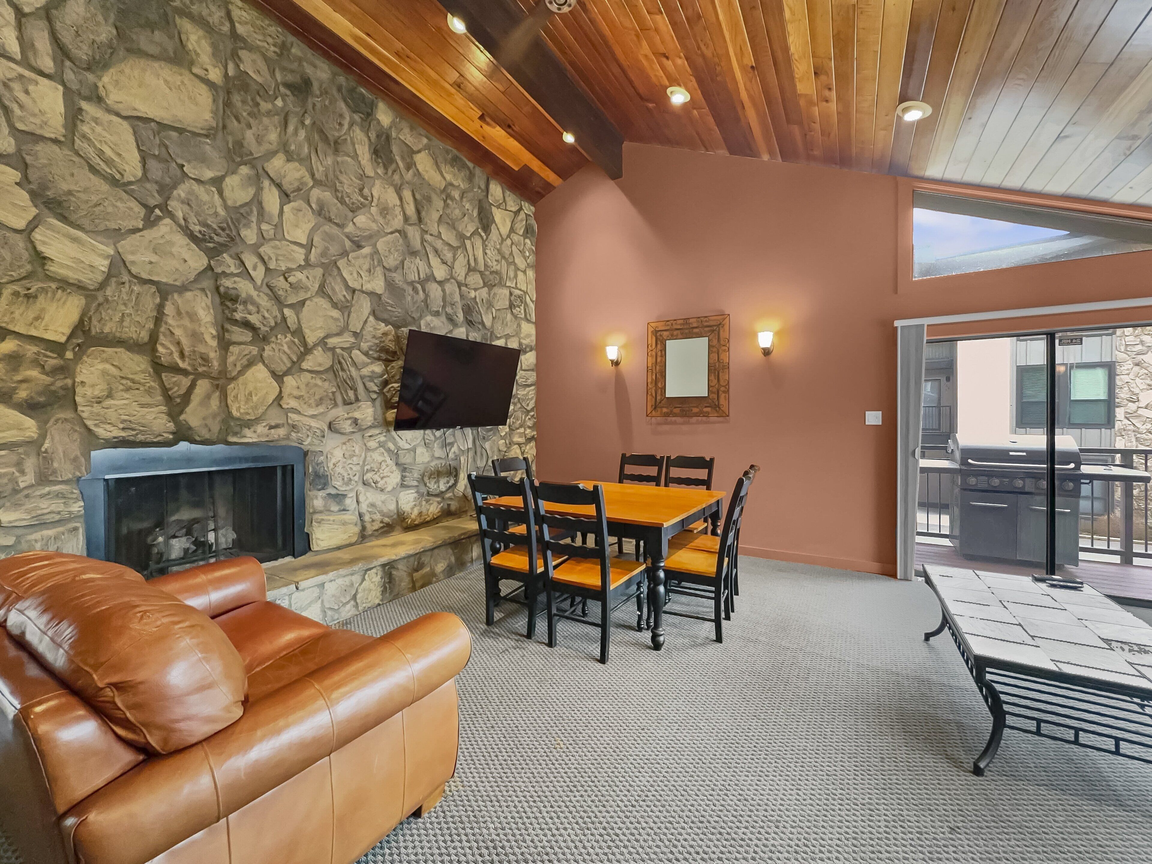A stone wall with a fireplace, a leather couch, a dining table with chairs, and a view of a barbecue grill outside.