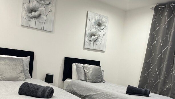 A hotel room with two beds, each with a black headboard, grey bedding, and a rolled grey towel. There are two wall-mounted paintings of flowers, and a grey curtain with a wavy pattern.